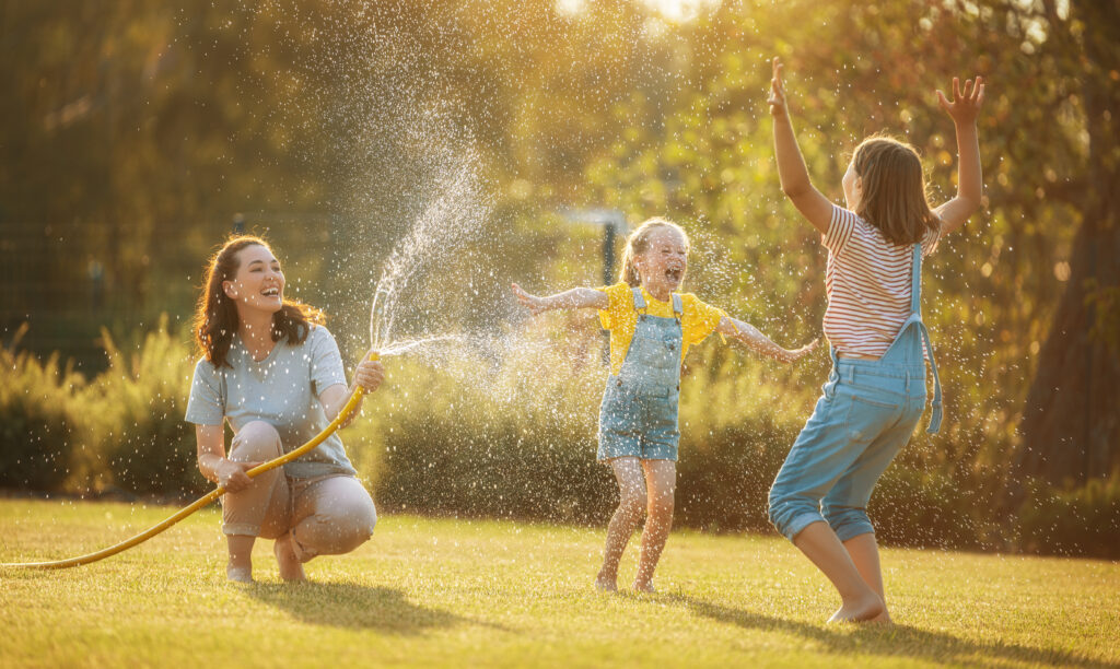 Happy family playing in backyard. Mother sprinkling her kids in hot summer day.