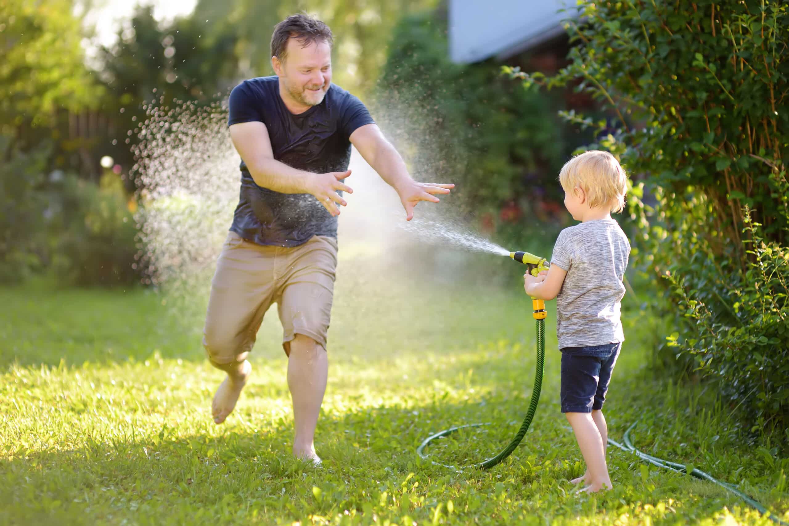 Funny little boy with his father playing with garden hose in sunny backyard. Preschooler child having fun with spray of water. Summer outdoors activity for kids.